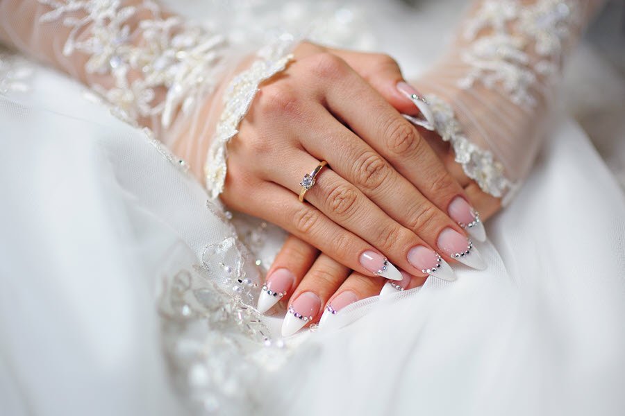 Bride showing a pale pink manicure with decorative nail accents while wearing a wedding dress.
