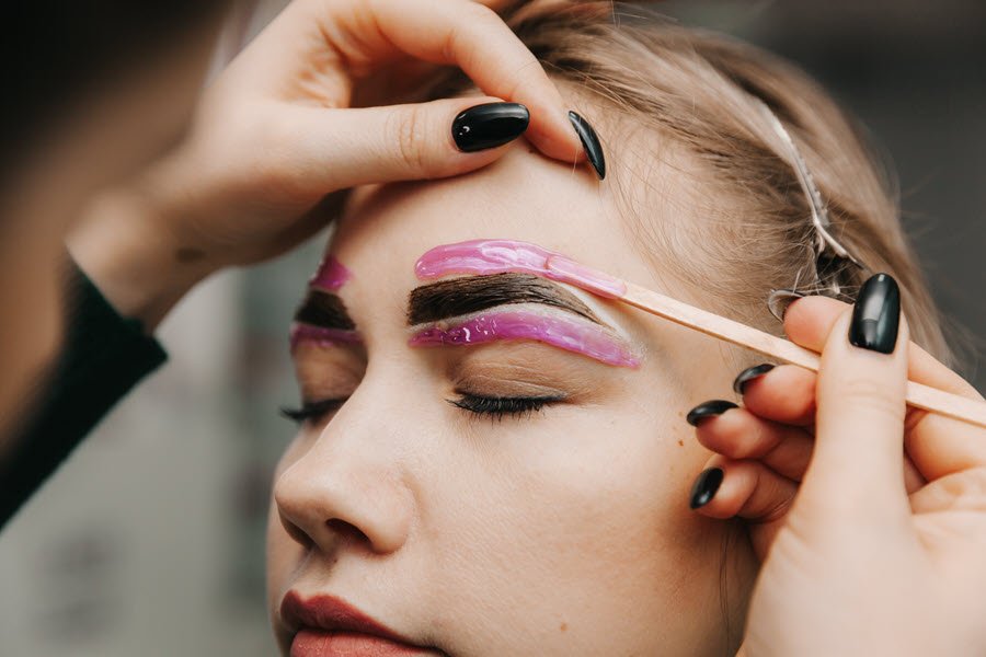 Beauty professional applying wax above a client’s eyebrow during a brow shaping service.