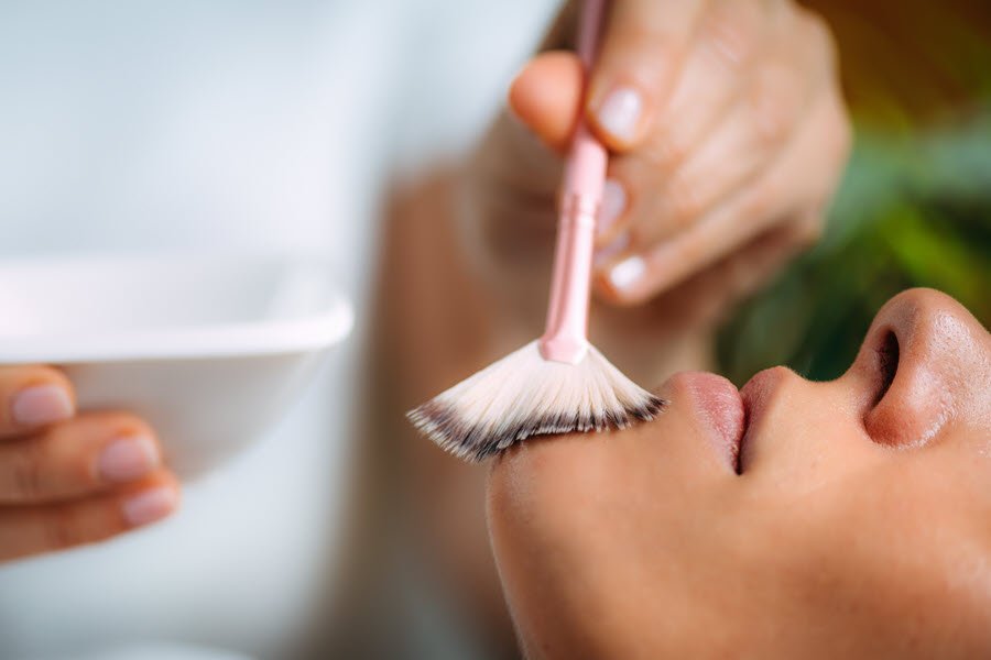 Esthetician applying a facial peel solution with a brush during a professional skin treatment.