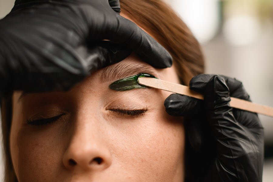 Beauty professional applying wax to a client’s eyebrow during a brow waxing service.