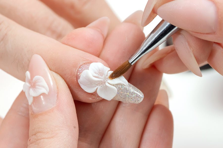 Nail artist creating a white 3D floral design with glitter accents on a client’s long nail.
