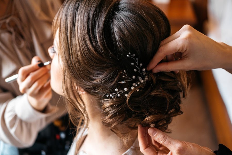 Stylist preparing a bride’s formal hairstyle while another beauty professional works nearby.